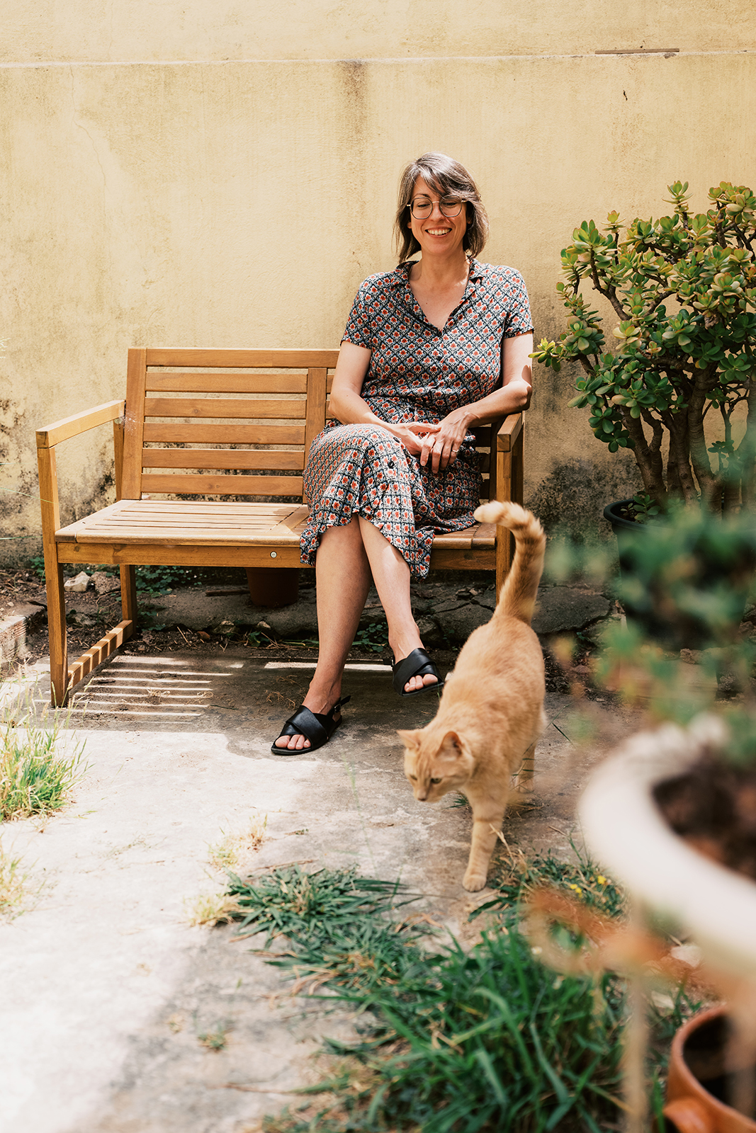 A photo Margaret sitting on a bench in her garden. In front of her walks her red cat Nelson.
